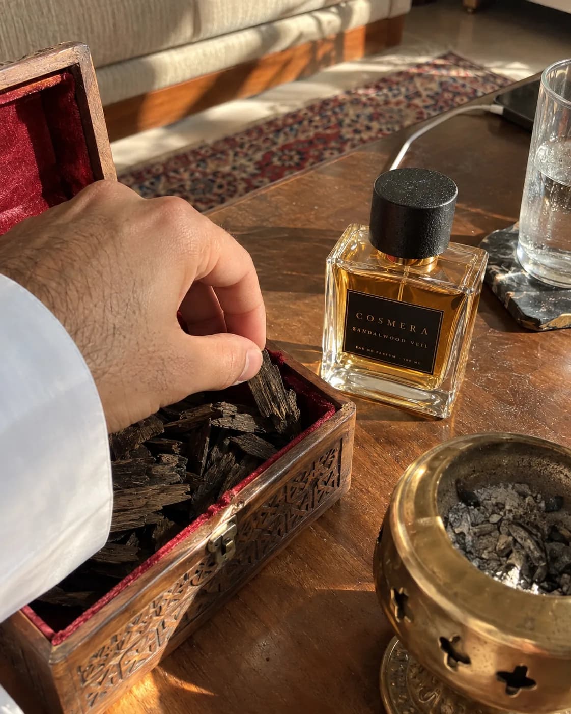 A hand in a white kandura sleeve picking a chip of oud from a carved wooden box, Sandalwood Veil and a brass mabkhara beside it