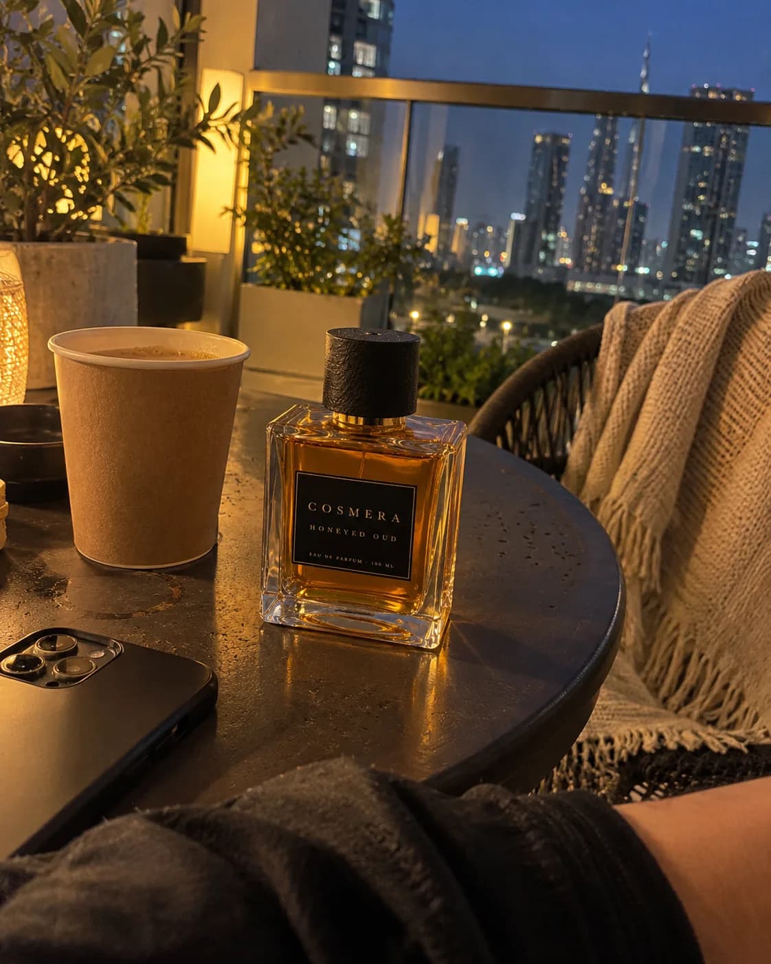Honeyed Oud on a small balcony table beside a cup of karak tea, a knitted throw on the chair and the Dubai skyline at dusk