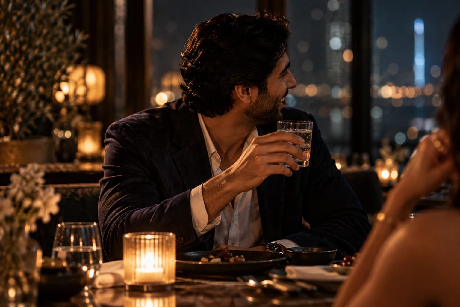 A man laughing over a late candlelit dinner, city lights behind him